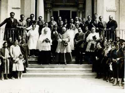 Per Goccia di Latte in quarant'anni di attivit� non mancarono onorificenze, premi e medaglie. Ecco una foto di una cerimonia tenutasi nel 1932, in occasione della visita all�ente dell�arcivescovo Augusto Curi. Si vedono volontari e sostenitori della Goccia posare sulle sue scalinate: Ave � in abito nero (e con il suo immancabile cappello con veletta) al centro tra il religioso e un altro parroco